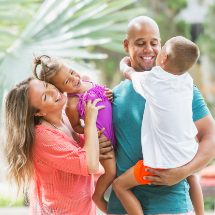 A man and a woman with two small children embrace in front of palm trees.