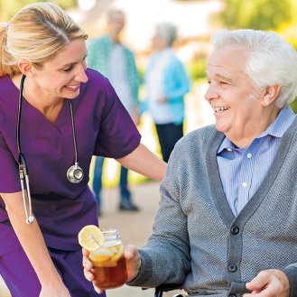 Elderly man accepts a glass of tea from nurse while sitting outside.